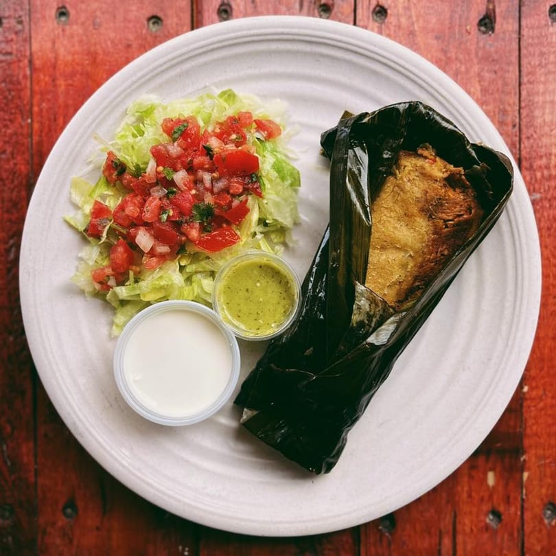 El Vaquero Mexican Taqueria tamale wrapped in a dark leaf, with lettuce, pico de gallo, and sauces.