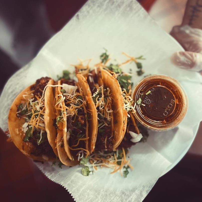 Four El Vaquero Mexican Taqueria tacos with cheese and cilantro, with a side of sauce.